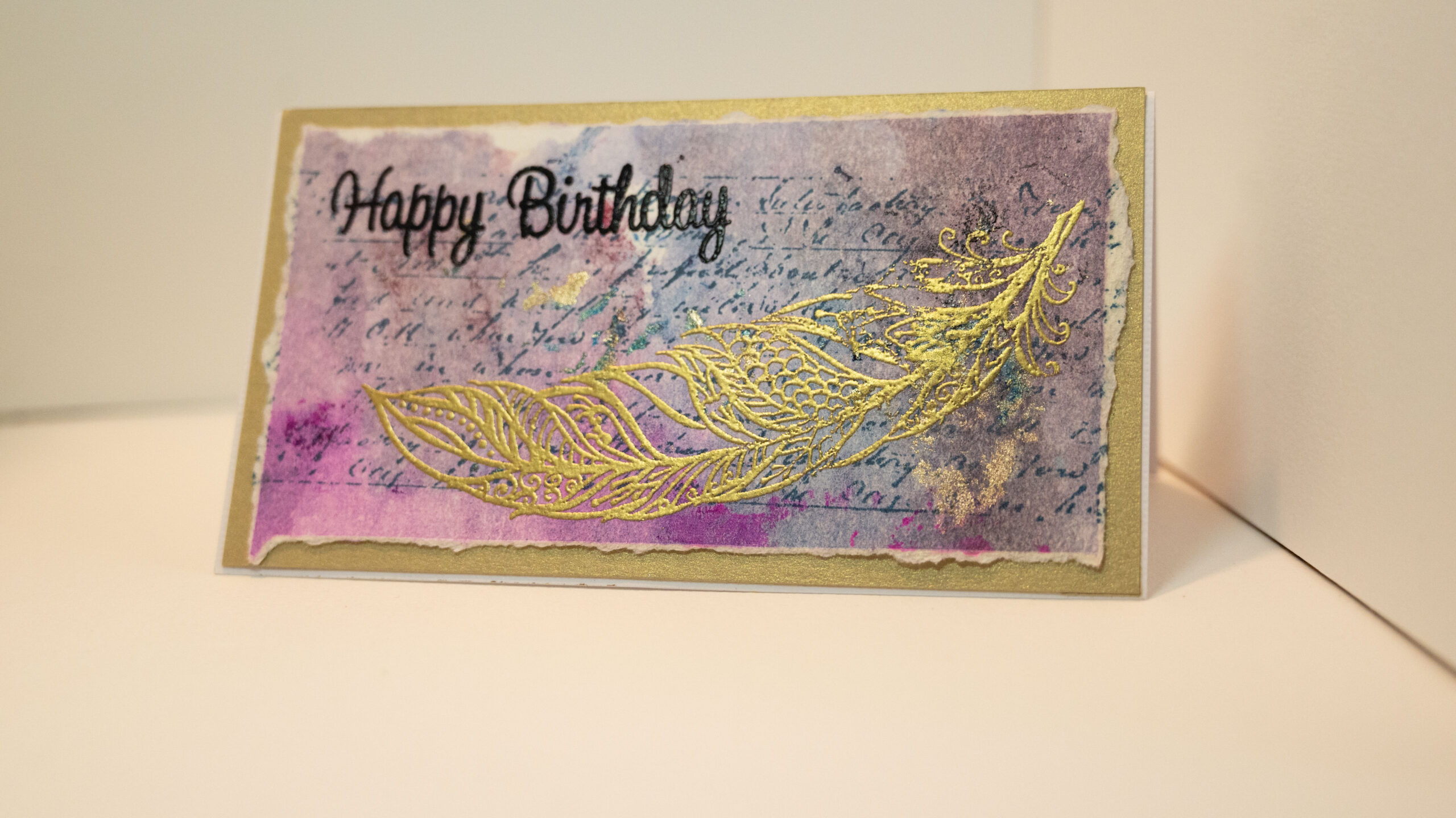 Hand made birthday card in pink, blue and gold, featuring a gold embossed feather outline and black embossed Happy Birthday sentiment. Mounted on hand coloured recycled paper.