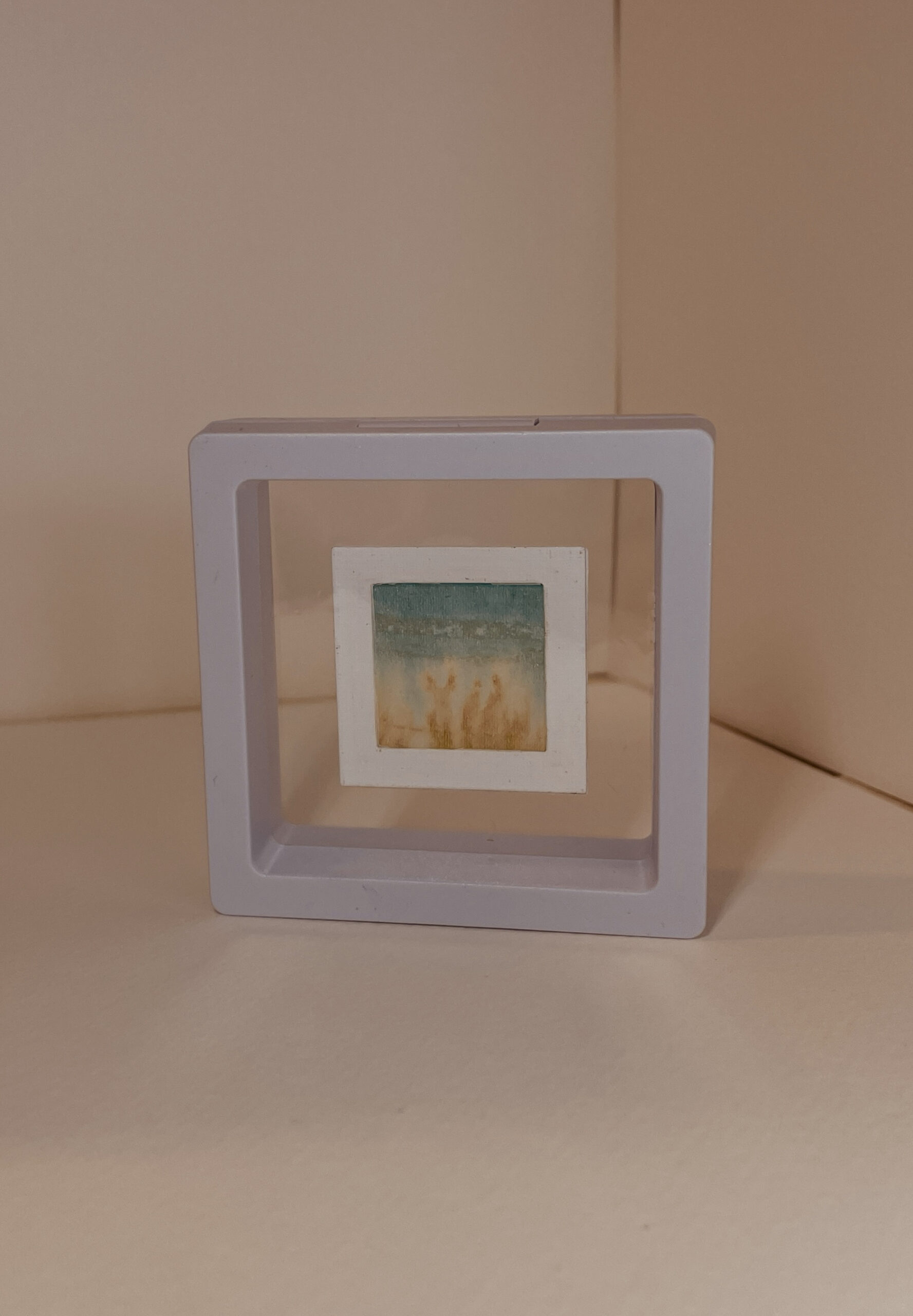 Reverse view of miniature landscape painted by hand on delicate tissue paper which is mounted within a card frame and within a transparent plastic click-frame. Gentle green and teal inks.