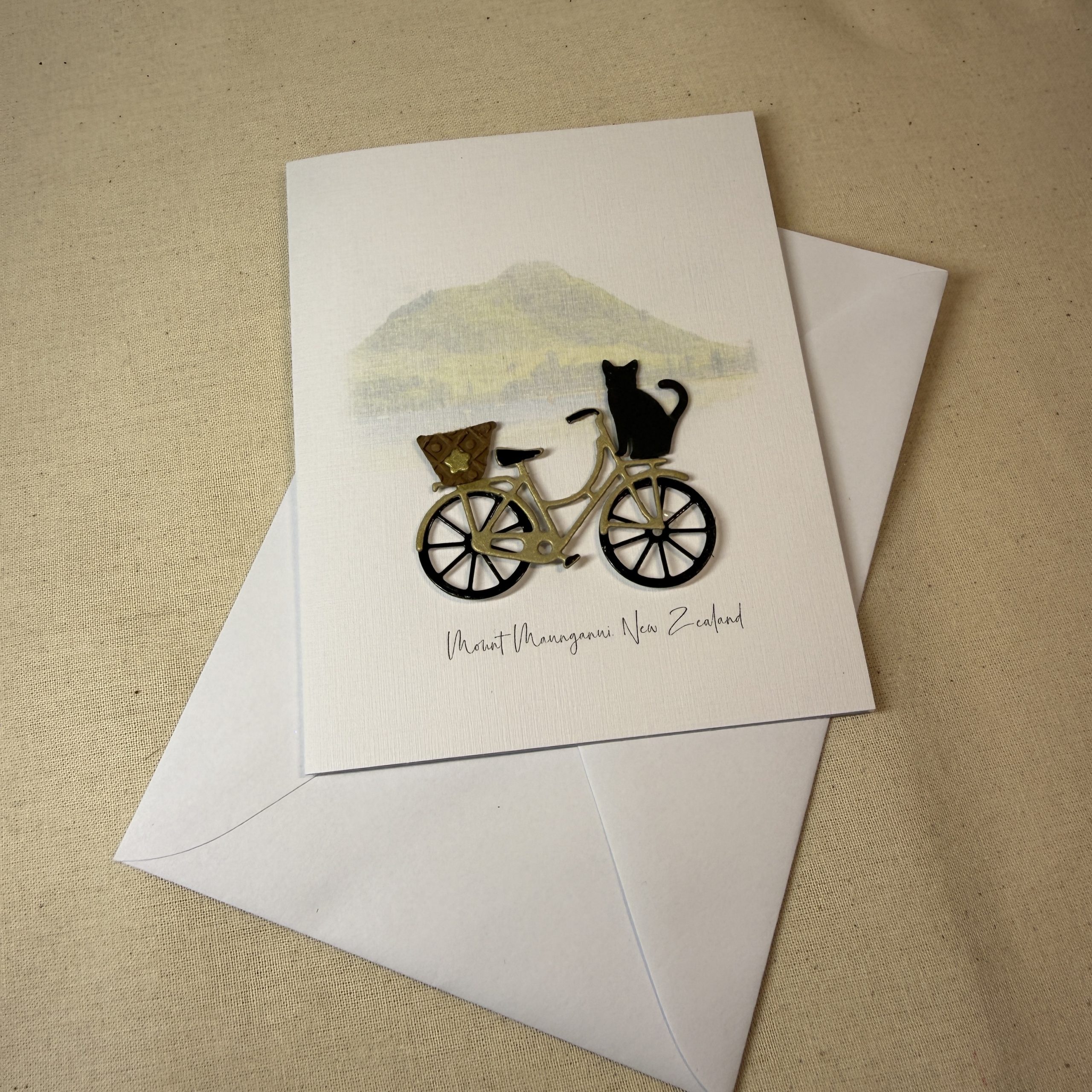 Mount Maunganui greeting card featuring a hand made bicycle with a basket and a black cat